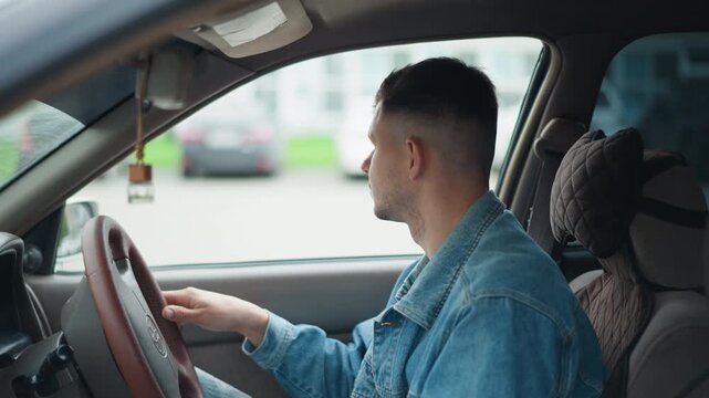 alert white driver scanning parking lot in denim jacket, hand on steering wheel while checking mirrors and blindside, prepares to reverse or merge, city background with parked cars, attentive posture