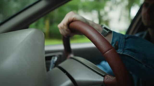 man gripping brown leather steering wheel inside car, trees and parkway visible through window denim sleeve and wristwatch in frame, closeup on hand executing smooth turn confident posture,