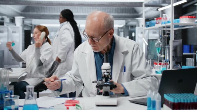 Histology technician analyzing bacterial culture slide to test antibiotic efficacy. Elderly man observing reaction under lens to develop new medical treatments, taking notes, camera B
