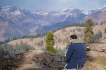 Man sitting on a mountain ledge overlooking a vast alpine valley and snow-capped peaks in the distance.
