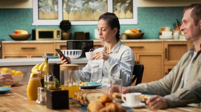 Preoccupied mother being detached and ignoring her family at the table, being distracted with a phone dependency and obsession during breakfast. Social media addiction. Camera B.