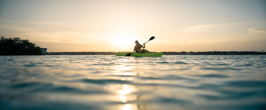 Silueta a contraluz de una hermosa mujer joven latina navegando en kayak en el mar, al atardecer, con el sol reflej&aacute;ndose sobre el agua