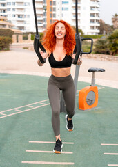 Strong redhead woman in sportswear exercising with suspension straps at outdoor gym during morning workout, maintaining active lifestyle and fitness routine