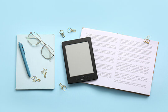Modern e-reader with book, notebook and eyeglasses on blue background
