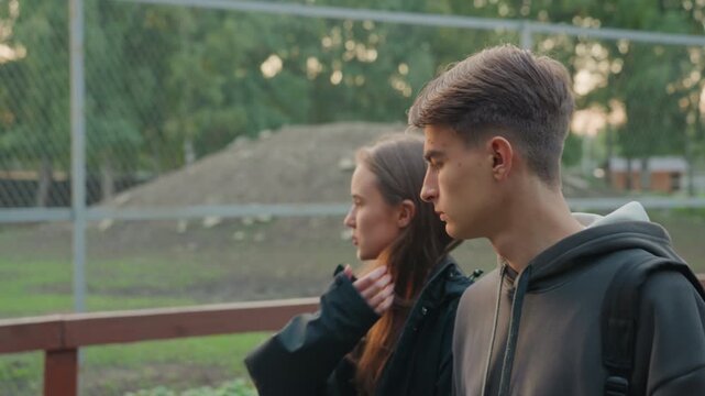 white teens pausing by fence together, girl tucking hair while boy watches with thoughtful expression against chain link and baseball field backdrop subtle gestures, listening and stolen glances