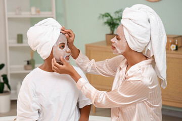 Happy young African-American mother and her daughter in pajamas applying facial sheet masks at home