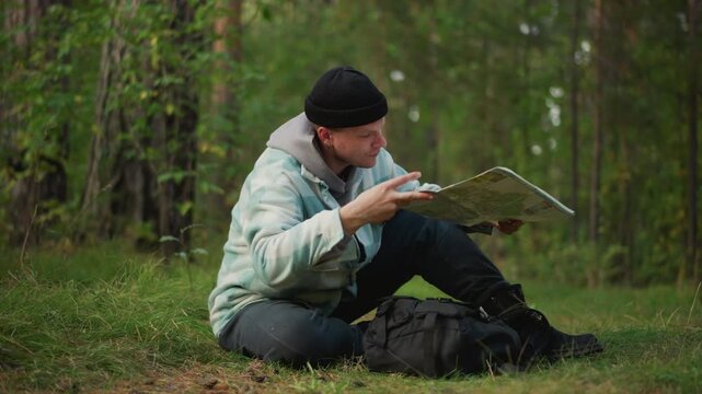 Explorer unfolding map in pine forest to locate trail and landmarks hopeful outdoors traveler inspects folds, adjusts position, reads chart against trees and moss, moment of clarity and satisfaction.
