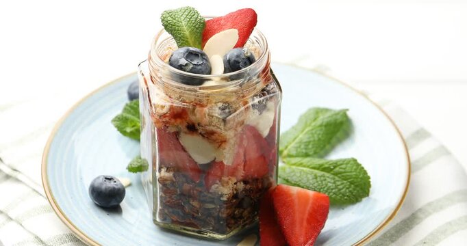 Tasty trifle dessert with berries, granola and mint in glass jar on white table, rotating