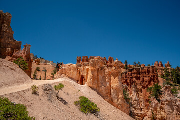 A rocky mountain with a blue sky in the background. The mountain is covered in trees and has a path leading up to it