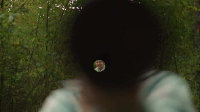 close barrel view shows man aiming, dark muzzle frames tiny focused reticle at center, blurred beanie and shoulders in foreground, dense green trees and soft bokeh background, scout inspects scope
