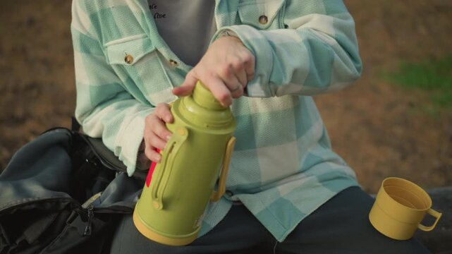 Male adventurer packs thermos into backpack after coffee break on trail, secures lid, stows cup into pocket and zips bag, preparing to resume hike through sunlit woodland with purposeful, practical