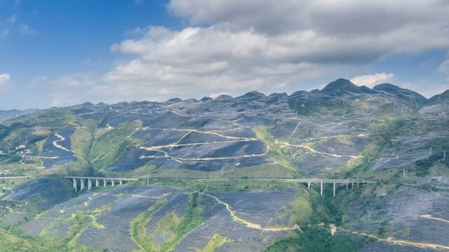 Aerial time-lapse of a massive mountain solar farm with highways cutting through vast photovoltaic arrays under a dynamic sky.