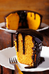 A slice of Brazilian carrot cake with a glossy, dripping chocolate frosting, served on a white plate with a rustic, blurred background