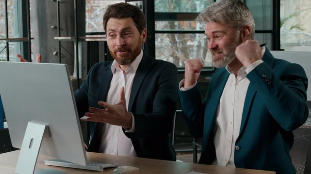 Two surprised happy Caucasian businessmen in office looking computer celebrating men business partner males guys employees colleagues pc screen victory win achieve successful deal high sales growth