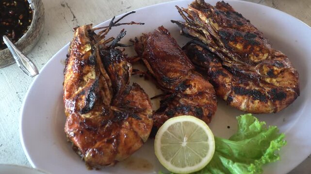 Delicious grilled giant tiger prawns served on a white plate with a slice of lemon and fresh lettuce. Close-up of Indonesian-style seafood BBQ with traditional sambal sauce on the side.