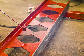 Red metal beam with hexagonal cutouts is being painted on concrete floor, showcasing industrial craftsmanship. vibrant color contrasts with industrial setting, highlighting work in progress