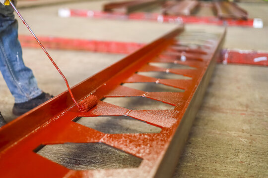 Worker is painting large metal beam with red paint, showcasing industrial craftsmanship. beam features lattice design, and scene captures focus on detail and precision