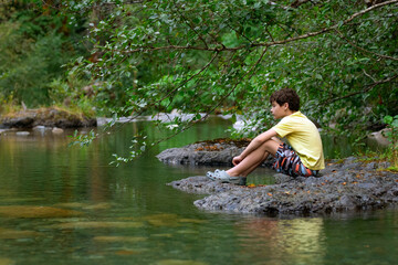 Fototapeta premium Teenager boy is sitting on a big rock in the mountain rocky river with clear green waters in summer forest. Scene under branches with green lush foliag