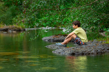Fototapeta premium Teenager boy is sitting on a big rock in the mountain rocky river with clear green waters in summer forest. Scene under branches with green lush foliag