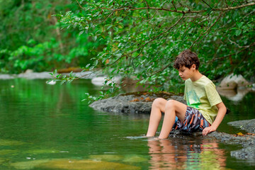 Fototapeta premium Teenager boy is sitting on a big rock in the mountain rocky river with clear green waters in summer forest. Scene under branches with green lush foliag