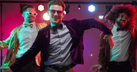 Dancers group perform disco dance under party lights. Three men in retro costumes wear glasses and bowties, dancing on stage for nightlife and ads. Energetic party and celebration concept.