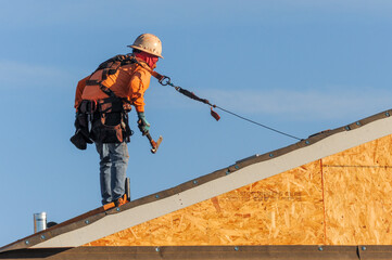 Construction worker installing roof underlayment on a two story home under construction, wearing fall prevention gear, representing residential roofing installation and exterior building operations