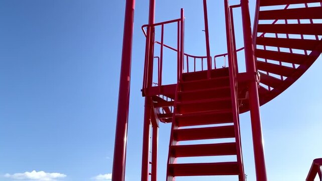 Red metal spiral staircase ascends into a bright blue sky with hints of white clouds