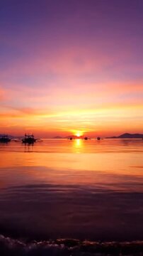 Calm ocean reflecting dramatic orange, pink, and purple sunset with silhouetted outriggers