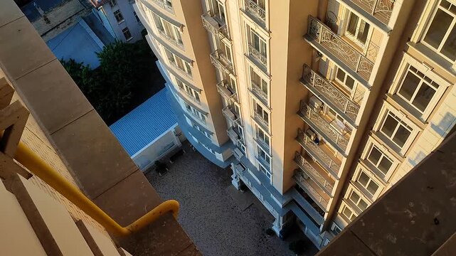 View down from top floor of multi-story building. Facade of white multi-story high-rise building with windows and balconies illuminated by light of setting sun and floor area underneath paved tiles