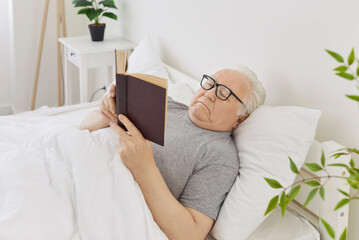 Portrait of gray-haired senior elderly man in eyeglasses reading a book lying on bed in his bedroom...