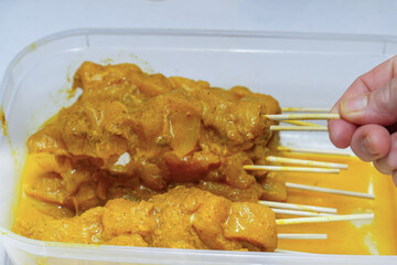 Hand placing raw chicken pieces on wooden skewers, immersing them in a yellow, turmeric-infused curry marinade within a clear container