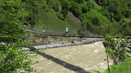 Blonde woman walking on a wooden suspension bridge over a forest river. A girl in a white tshirt and gray pants crosses the hanging bridge surrounded by trees and flowing water.