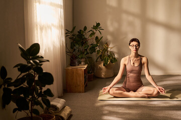Caucasian young adult woman sitting cross legged on yoga mat meditating with eyes closed in bright...