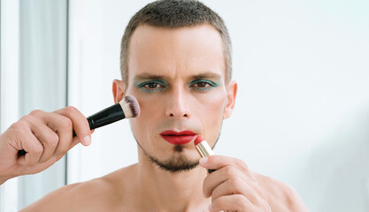 gay man with makeup and cosmetics on face. transgender man with bright eyeshadows paints lips with red lipstick and applies a blush brush to his face on a white background