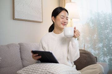 Relaxed woman enjoying a cup of coffee while holding digital tablet on a comfortable sofa at home....