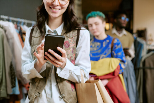 Young adult woman smiling while using smartphone and holding shopping bag in second hand clothing store, diverse young adults browsing racks in background