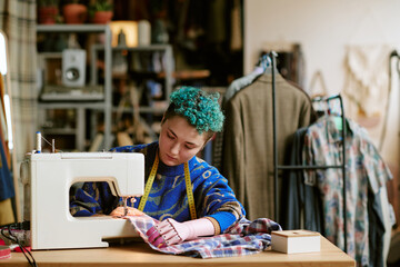Young adult Caucasian woman with prosthetic hand using sewing machine repairing plaid shirt in...