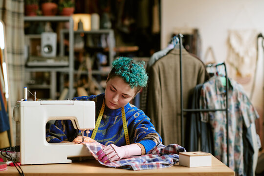 Young adult Caucasian woman with prosthetic hand using sewing machine repairing plaid shirt in second hand clothing store, measuring tape around neck, racks of clothes in background