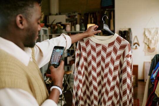 Young adult Black man photographing patterned sweater with smartphone in second hand clothing store, holding hanger and focusing on capturing garment details for online resale