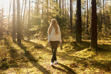 Woman walking through a sunlit forest path surrounded by trees and greenery in autumn