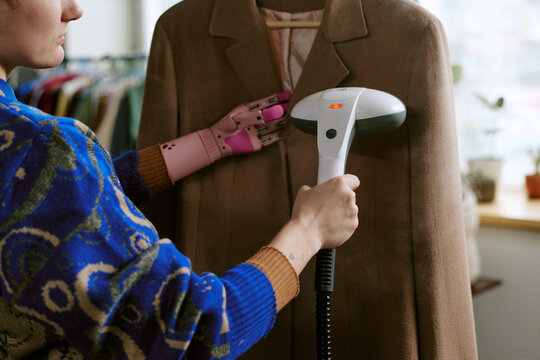 Young adult woman with prosthetic hand steaming second hand coat on hanger, focusing on garment care in resale or thrift store setting, clothing rack in background