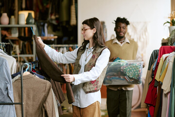 Biracial young adult woman browsing second hand clothing rack, while Black young adult man carrying plastic bin filled with assorted garments in thrift store environment
