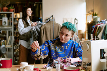 Young adult Caucasian woman with prosthetic hand sorting fabric scraps at table with sewing machine, middle aged Caucasian woman browsing second hand clothing rack in background