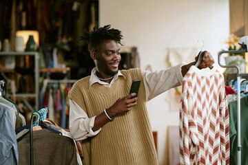 Young adult Black man smiling while holding smartphone and examining patterned dress on hanger in second hand clothing store, surrounded by racks of assorted garments