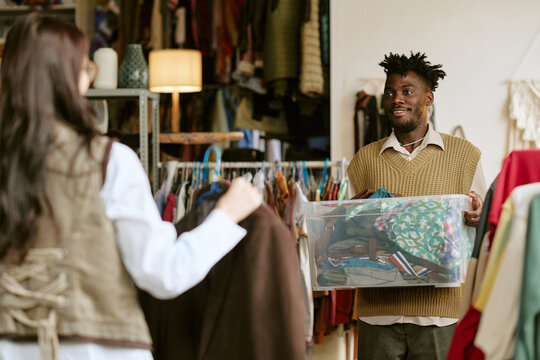 Young Black man holding plastic container filled with assorted second hand clothes, standing in thrift store while young woman examining garment in foreground