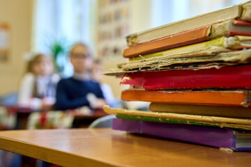 Stack of worn colorful books on a classroom desk with children blurred in the background, symbolizing learning, education, and everyday school life.