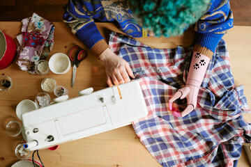 Young adult Caucasian woman with prosthetic arm sewing plaid dress using sewing machine on wooden table, surrounded by fabric, scissors, thread and beads, representing second hand clothing