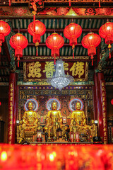 Fototapeta premium Golden Buddha statues inside Wat Mangkon Kamalawat, Bangkok Chinatown. Soft blurred worship candles in foreground and red lanterns above create a peaceful Lunar New Year spiritual atmosphere.