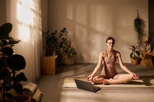 Caucasian young adult woman sitting cross legged on yoga mat meditating in bright room, using laptop for online yoga class, surrounded by indoor plants, wearing glasses