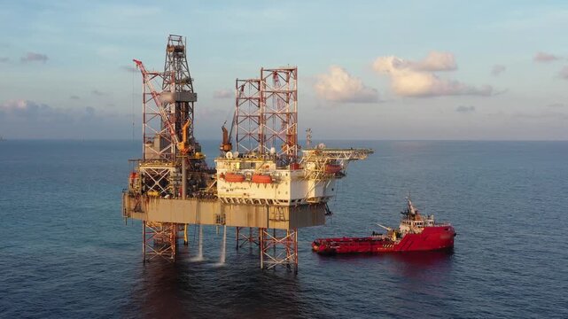 Aerial view from a drone of big jack up offshore drilling oil rig, production platform and supply vessel In the ocean during sunset - Oil and Gas Industry
